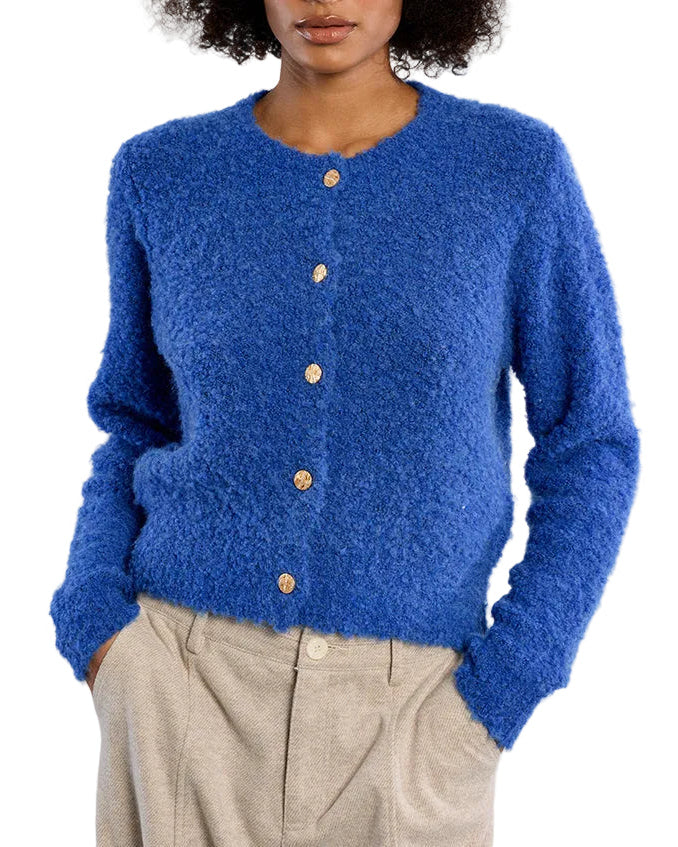 A woman with dark curly hair wearing a fuzzy royal blue button-up cardigan and beige trousers, standing against a light gray studio background.