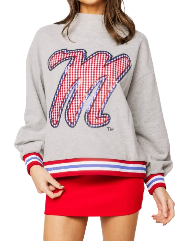 A model wearing a light gray sweatshirt with a large red and white gingham checkered 'M' graphic on the front, paired with a red mini skirt. The sweatshirt features red, white, and blue striped ribbed cuffs and hem.
