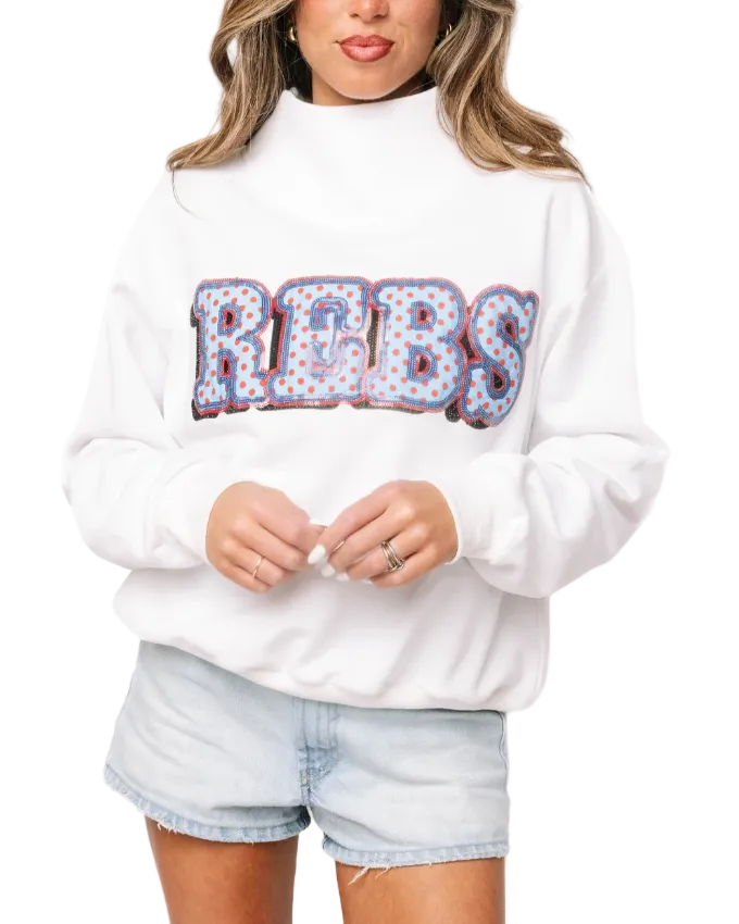 A woman wearing a white mock-neck sweatshirt with a large 'REBS' graphic across the chest in a patriotic red, white, and blue pattern, paired with light-wash denim shorts.