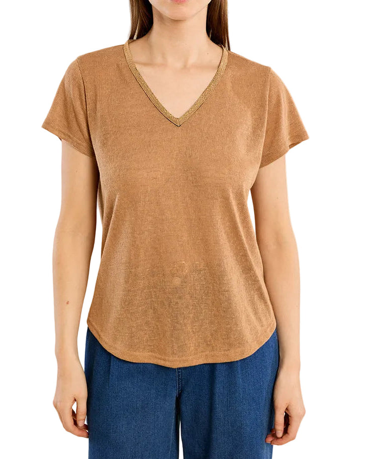 A woman modeling a short-sleeve V-neck top in a warm tan or camel color, paired with blue pants, against a solid light beige background.