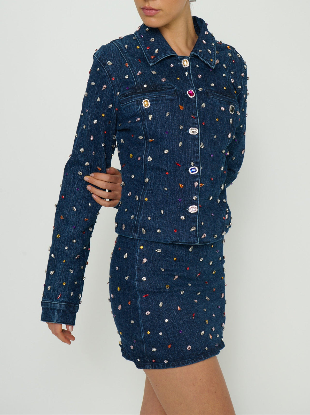 A model wearing a dark blue denim jacket and matching mini skirt set, both heavily embellished with small colorful embroidered motifs, against a plain light gray background.