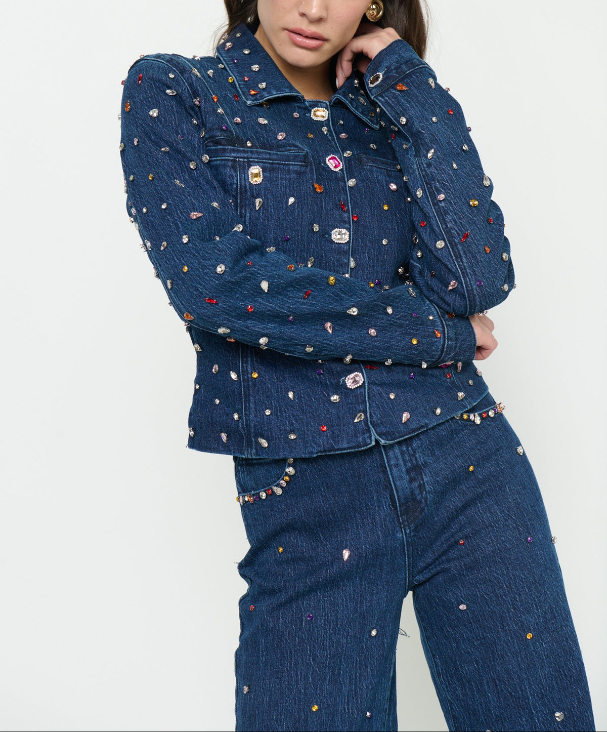 A model wearing a dark blue denim jacket and matching jeans, both heavily embellished with small colorful rhinestone studs, against a plain white studio background.